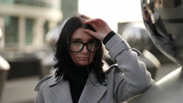 Portrait of a Brunette with Glasses and a Gray Coat Near Metal Structures on a Windy Day alt