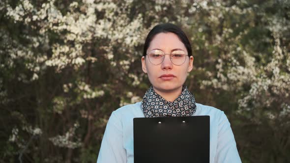 A Woman in Glasses with a Black Folder in Her Hands Looks at the Camera alt