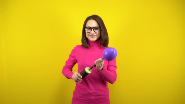 A Young Woman Inflates a Purple Balloon with a Pump on a Yellow Background. Girl in a Pink alt