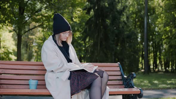 Young Girl Sitting on a Park Bench Wearing Coat and a Hat Writing in Notebook alt