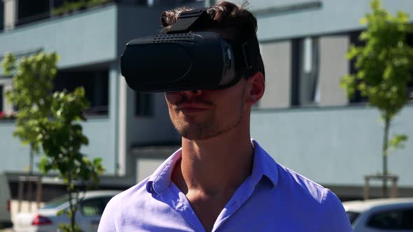 Young Handsome Man Uses Virtual Reality Glasses - Buildings in the Background - Closeup alt
