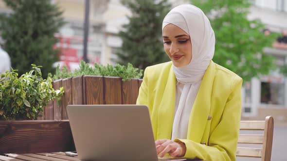 Smiling Young Muslim Woman Wearing Headscarf Sits in Cafe and Uses Laptop