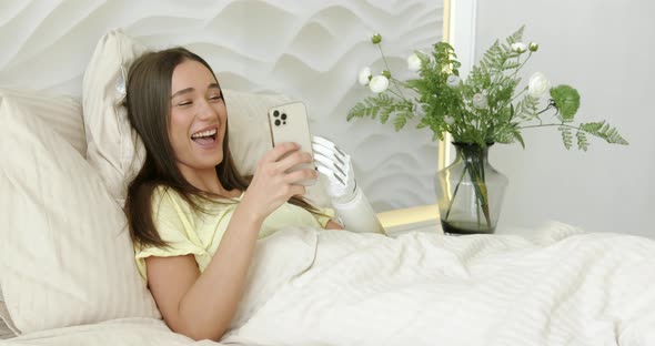 Girl with Bionic Arm Using Phone Reading Social Media and Chatting in Bed Woman with Disability alt