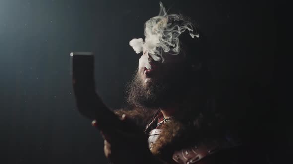 Bearded Man in Shaman Clothes Breathes Out Smoke on Black alt