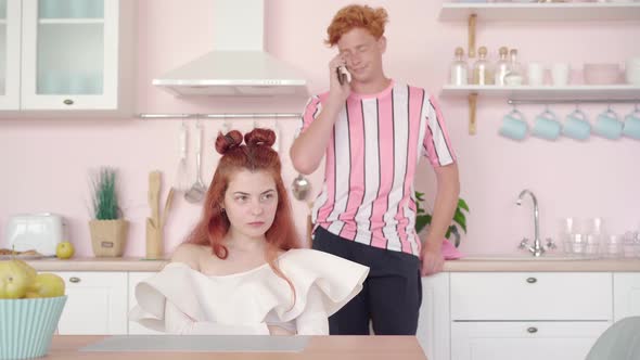 Unhappy Redhead Woman Sitting at the Table in Pink Kitchen As Man Talking on the Phone at the alt