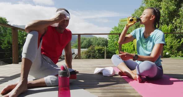 Tired biracial couple on terrace drinking water, resting after exercises and talking alt