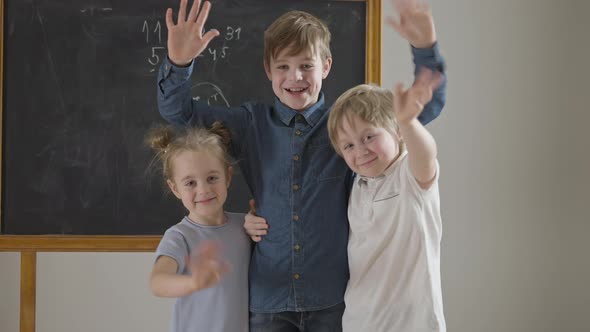 Cheerful Caucasian Friends or Siblings Waving Looking at Camera Standing in Classroom at Chalkboard alt