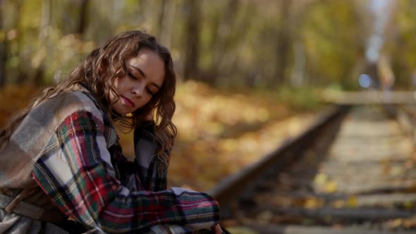 a Woman with Curly Hair and a Plaid Coat Sits Sadly on the Rails in an Autumn Park on a Sunny Day alt