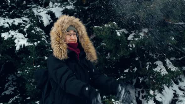 An Attractive Woman in a Green Hat and a Fluffy Hood Walks in a Snowcovered Park Throws Snow with