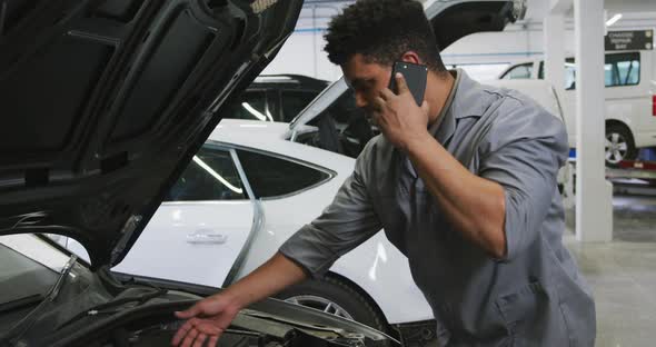 African American male car mechanic looking at an open car engine and talking on a smartphone alt