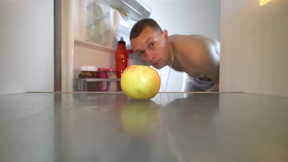 young man opens refrigerator and sees empty shelves. One apple is lying. alt