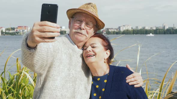Attractive happy elderly couple in 60s take selfie with smartphone at sunset on lake smiling and lau