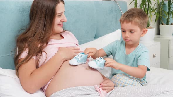 Smiling Pregnant Woman Looking on Her Little Son Playing with Baby Boots on Her Big Belly alt