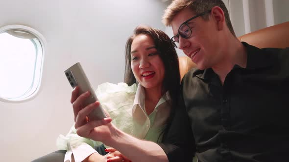 Happy couple passengers in love traveling see smartphones and smile together on the airplane