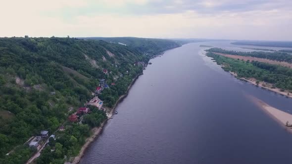 Camera is Flying Over Wide River Along Shore with Small Houses Aerial View of Landscape