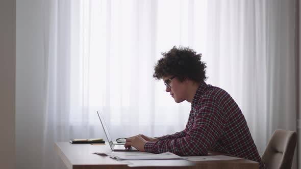 Curly Haired Male Student Attractive Young Boy in Glasses is Studying at Home Using Laptop Typing alt
