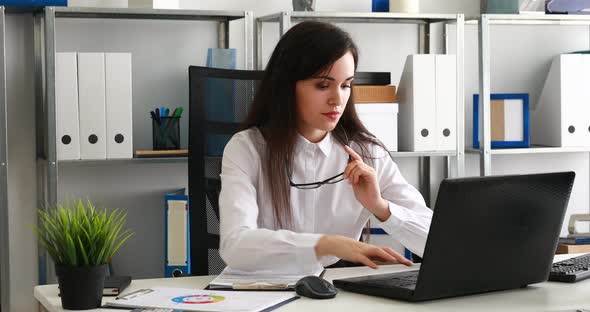 Businesswoman Taking Off Glasses and Looking Into Camera, Working on Laptop in Modern Office