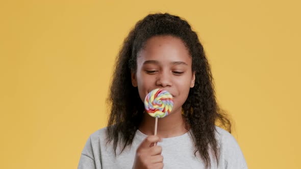 Studio Portrait of Positive African Teen Girl Licking Rainbow Lollipop Enjoying Sweets and Smiling alt