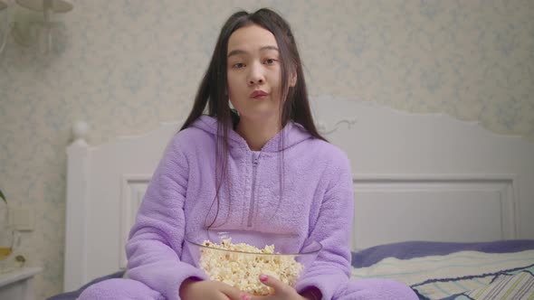 Bored 20s Asian Woman Watching TV and Eating Popcorn Sitting Alone on Bed alt