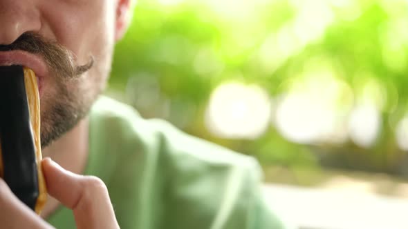 Bearded Man with Curled Up Mustache Eats Chocolate Eclair in Cafe. Half Faces in the Frame with alt