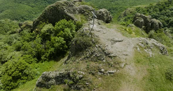Simple Christian cross on a rocky hill surrounded with thick, green bushes. alt