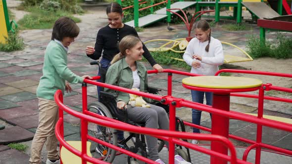 Cheerful Children Spinning Disabled Girl in Wheelchair on Merry Go Round Outdoors alt