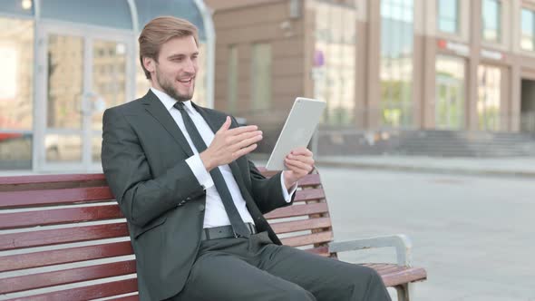 Online Video Chat on Tablet By Businessman Sitting Outdoor on Bench alt