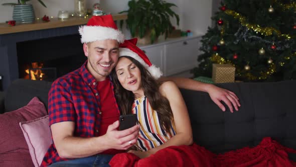 Happy caucasian couple wearing santa hats sitting on sofa and using smartphone alt