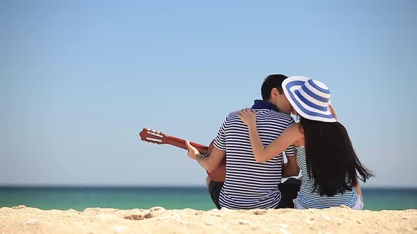 Young couple with guitar sits on sand on the beach in summer day.