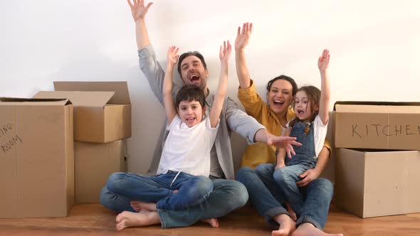 Happy Multiracial Family of Four Sitting on Floor Surrounded Cardboard Boxes in Empty Living Room alt