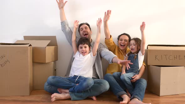 Happy Multiracial Family of Four Sitting on Floor Surrounded Cardboard Boxes in Empty Living Room