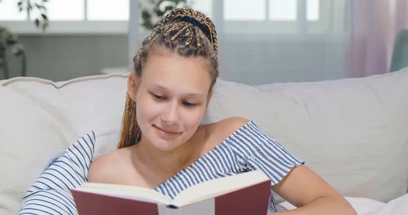 Close-up Shot of Young Attractive Teenage Girl with Breadlocks Lying on Sofa and Reading Book alt