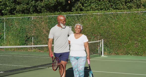 African american senior couple holding rackets walking on the tennis court alt