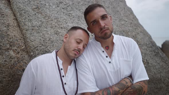 Gay Couple with Closed Eyes Resting Leaning on Each Other Shoulder By the Rock Looking at the Camera alt