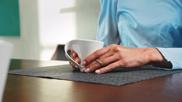 Hand Holds a White Mug with Coffee While Sitting at a Table in the Living Room alt