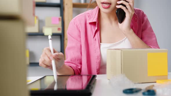 Young Asian woman using phone call receiving purchase order and check product on stock work at home. alt