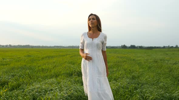 Beautiful Young Woman in White Dress Walks on the Green Field. Wind Blowing Hair