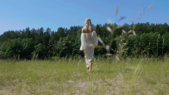 Blonde Girl in White Flowing Dress Walks Barefoot on Grass with Sense of Oneness with Nature