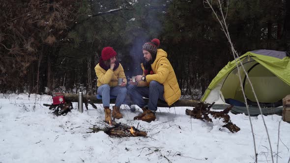 Young Man and Woman Drink Tea By the Fire in the Winter Forest alt