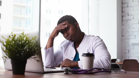 Tired African Black Male Doctor Working on Laptop, Thinking About Problem and Drinking Coffee. alt
