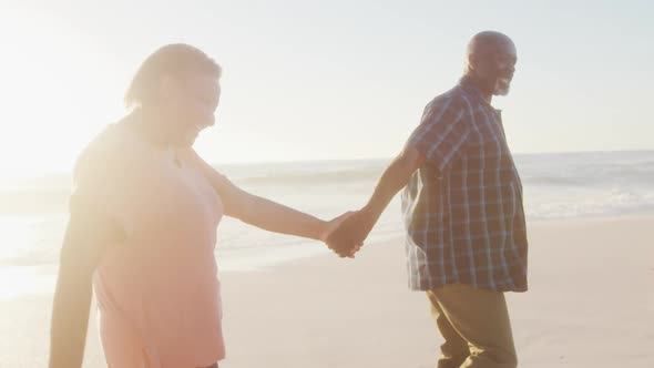 Smiling senior african american couple holding hands and walking on sunny beach alt