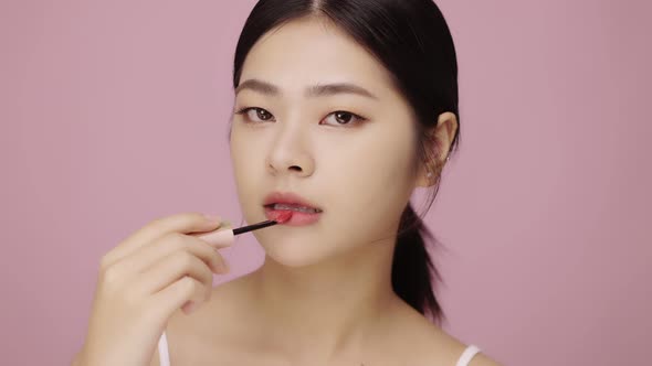 Close up face of beautiful young Asian woman applying lip on her lips on pink background. alt