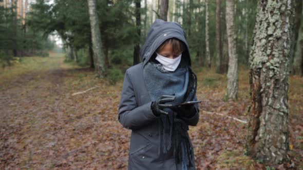 A Girl with a Face Mask Uses a Smartphone Alone in the Woods