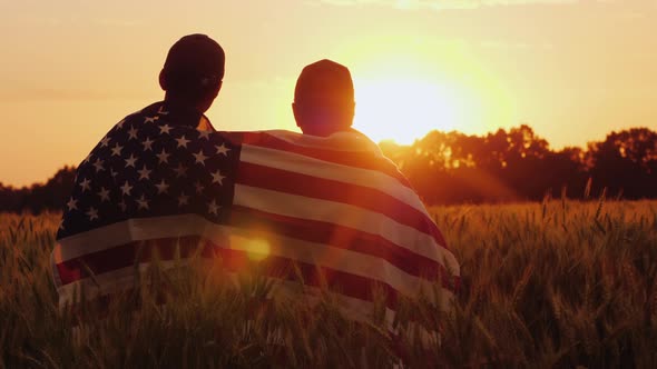 A Man and His Son Admire the Sunset Over a Field of Wheat, Wrapped in the Flag of the USA alt