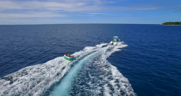 Aerial drone view of man and woman on an inflatable tube towing behind a boat to a tropical island. alt