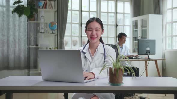 Young Asian Woman Doctor Is Using Laptop Computer And Smiling To Camera In Workplace alt