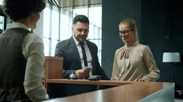 Young Couple Checkingin Hotel Talking To Receptionist Paying for Room with Smartphone Getting Key alt
