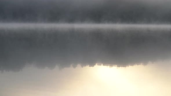 Morning Fog Blows Slowly Across a Clam Lake with Trees Reflecting in the Water