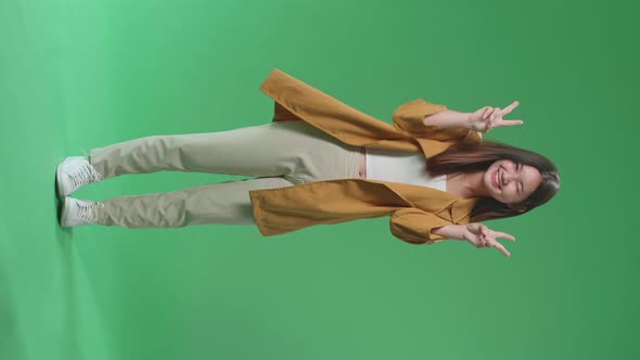 Full Body Of Smiling Asian Woman Showing Gesture Peace While Standing On Green Screen In The Studio alt