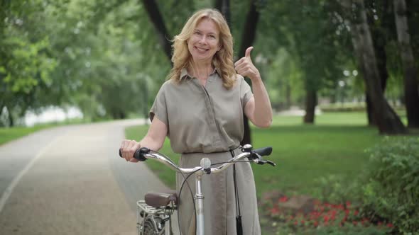 Portrait of Joyful Blond Middle Aged Woman Standing with Bike Looking at Camera Smiling and Showing alt